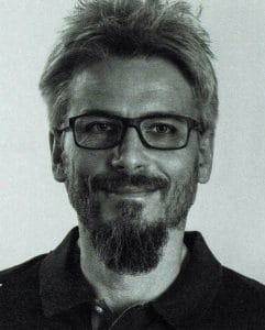 Ein Schwarzweißfoto eines Mannes mit kurzen Haaren, Brille und Bart. Er lächelt leicht und trägt ein dunkles Hemd mit Kragen vor einem schlichten Hintergrund.