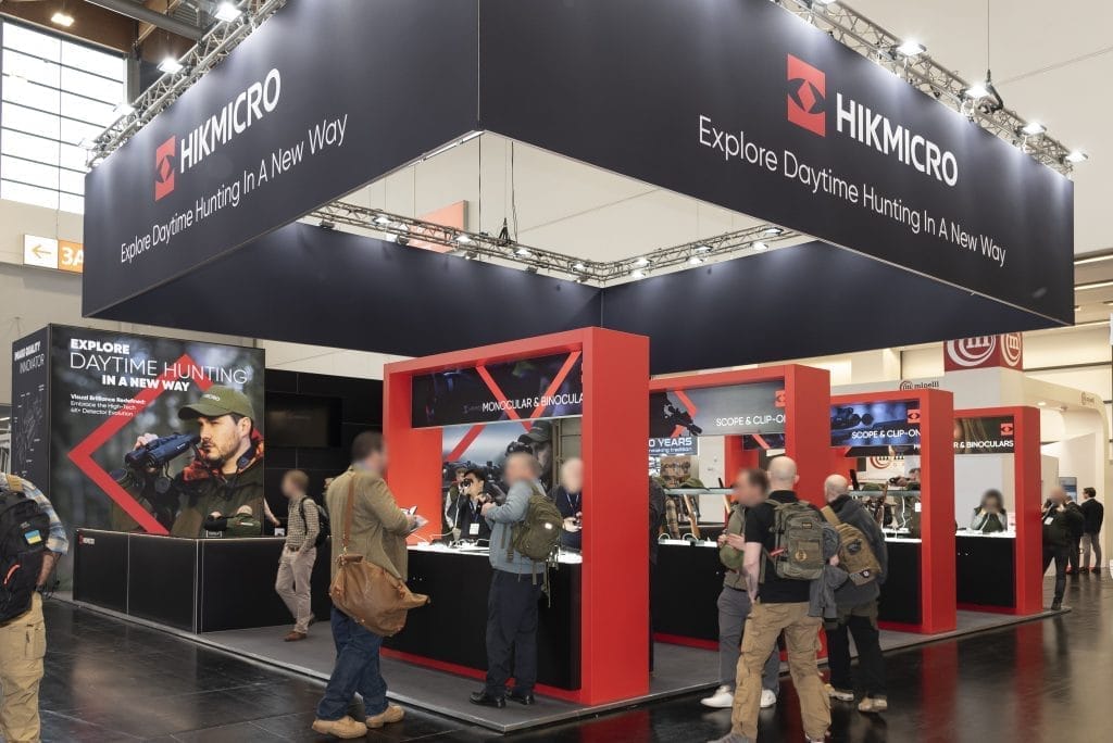 Messestand mit dem Branding „HIKMICRO“. Besucher stöbern in der Ausstellung mit Jagdausrüstung. Der Stand ist mit roten und schwarzen Akzenten und Bannern mit der Aufschrift „Entdecken Sie die Tagesjagd auf neue Weise“ versehen. Verschiedene Produkte werden auf Theken präsentiert.