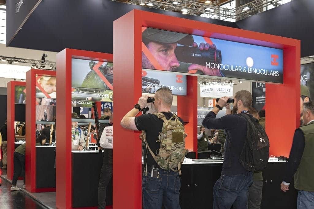 Zwei Personen in Freizeitkleidung und mit Rucksäcken benutzen Ferngläser an einem Messestand mit rot umrahmten Displays. Der Stand präsentiert monokulare und binokulare Produkte. Im Hintergrund sind weitere Besucher zu sehen.