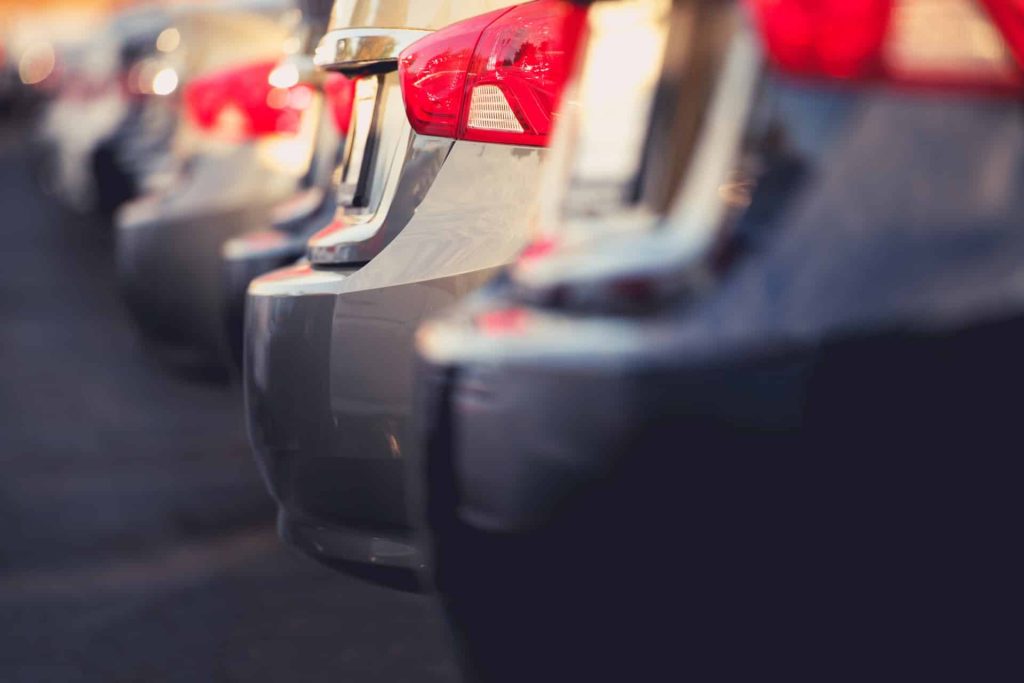 Eine Nahaufnahme zeigt geparkte Autos in einer Reihe, deren Heckstoßstangen und Rücklichter leuchten. Die Aufnahme aus einem Winkel, um Tiefe und Ausrichtung zu erzeugen, erinnert an die geordnete Anordnung des größten Messegeländes Deutschlands, wobei die Beleuchtung einen hellen, sonnigen Tag suggeriert.