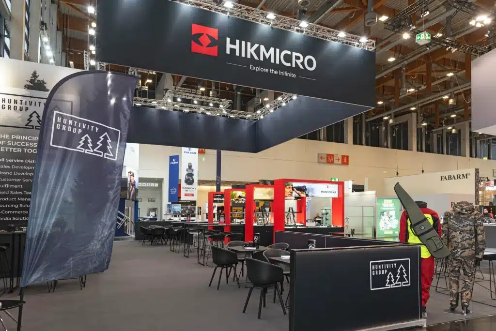 Messestand mit Hikmicro-Schildern und dem Slogan „Explore the Infinite“. Der Stand umfasst verschiedene Produktpräsentationen, Sitzbereiche mit Tischen und Stühlen sowie ein Banner der Hunttivity Group. Die Location ist eine große, gut beleuchtete Ausstellungshalle.