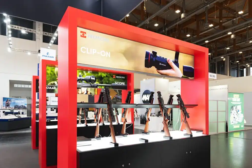 Ein Messestand mit roten Rahmen zeigt Gewehre in Halterungen. Ein Banner darüber zeigt Hände, die ein Clip-On-Gerät halten. Der Stand präsentiert den Markennamen „HIKMICRO“ und Schilder für Produkte wie „Clip-On“ und „Scope“.