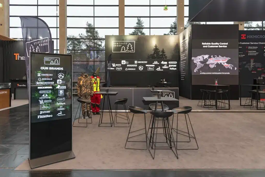 Messestand mit Stühlen, Tischen und Markendisplays. Auf einem Bildschirm werden verschiedene Logos und Markennamen präsentiert. Dahinter zeigen eine große Karte und Text die weltweiten Servicegebiete. Eine Person in Warnkleidung steht daneben.