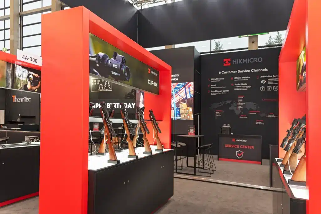 Messestand mit Gewehren und Zielfernrohren. Rot-schwarze Displays mit verschiedenen Produkten und Informationsschildern. Große Banner präsentieren Markenlogos und Kundenserviceinformationen. Hell beleuchteter Ausstellungsbereich mit modernen Designelementen.
