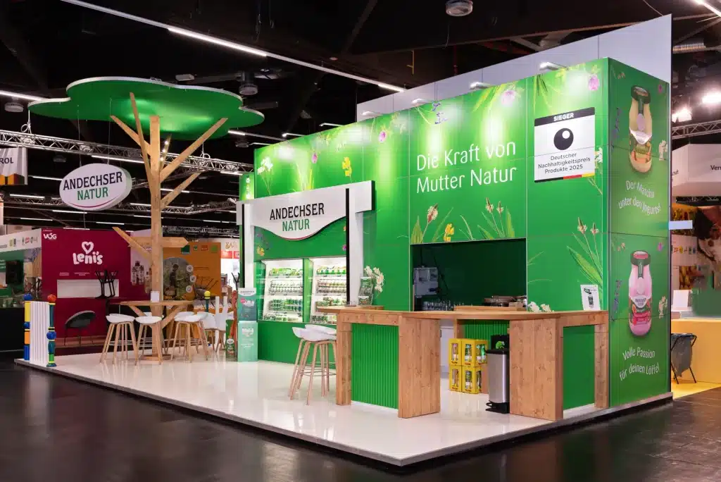 Grüner Messestand von Andechser Natur in Nürnberg mit Slogan Die Kraft von Mutter Natur, Holzbaum, Theke und beleuchteter Produktwand mit Molkereiprodukten.