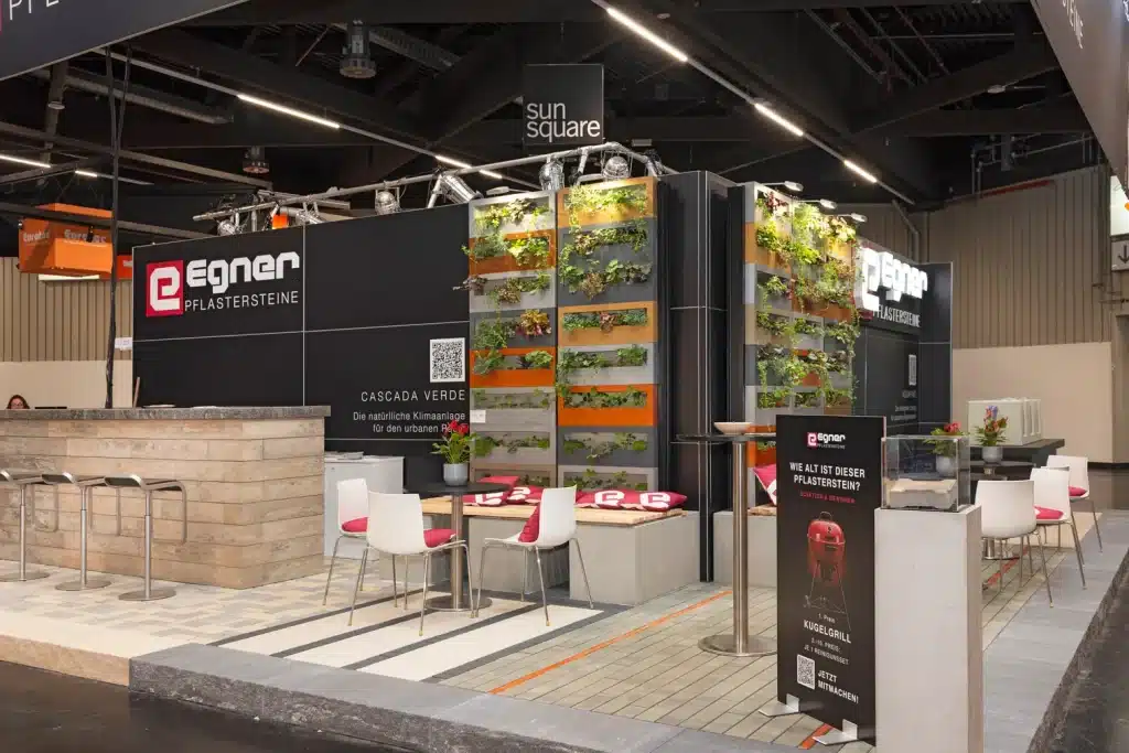 Messestand von Egner Pflastersteine auf der GaLaBau in Nürnberg mit umlaufendem schwarzem Logoblender, Pflasterfläche und Begrünungswand.