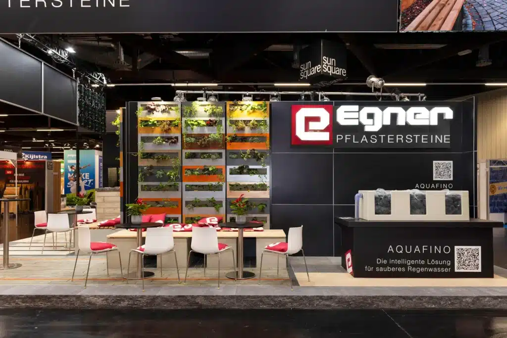 Egner-Messestand auf der GaLaBau in Nürnberg mit langer Tafel, roten Polsterstühlen und beleuchteter Pflanzenwand in Orange und Grün.