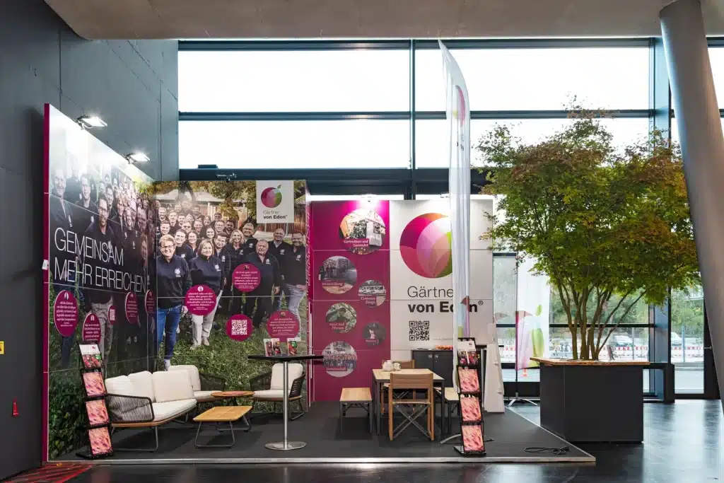Messestand von Gärtner von Eden auf der GaLaBau in Nürnberg mit Teamfoto-Wand, weißer Lounge, Beachflag und Ahorn im Pflanzkübel.