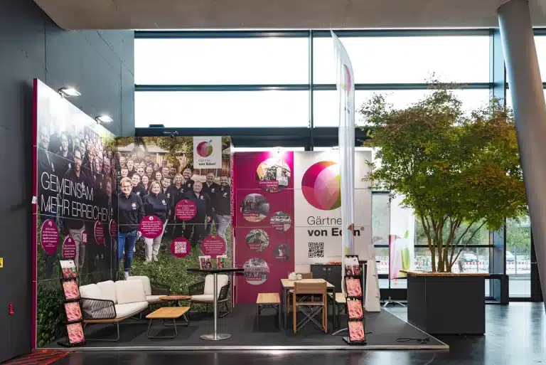 Messestand von Gärtner von Eden auf der GaLaBau in Nürnberg mit Teamfoto-Wand, weißer Lounge, Beachflag und Ahorn im Pflanzkübel.