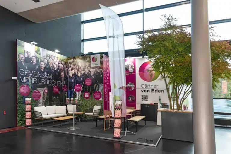 Messebau für Gärtner von Eden auf der GaLaBau Nürnberg mit magentafarbener Logowand, Sitzgruppe und Banner Gemeinsam mehr erreichen.
