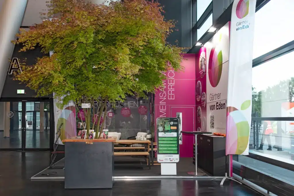 Seitenansicht des Stands von Gärtner von Eden auf der GaLaBau in Nürnberg mit großem Ahorn, Infoterminal und Beachflag vor der Hallenfront.