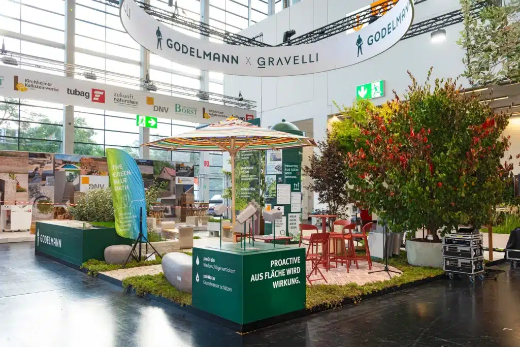 Messebau für Godelmann auf der GaLaBau in Nürnberg mit Sonnenschirm, roter Sitzgruppe, Rasenfläche und grünen Sockelwänden.