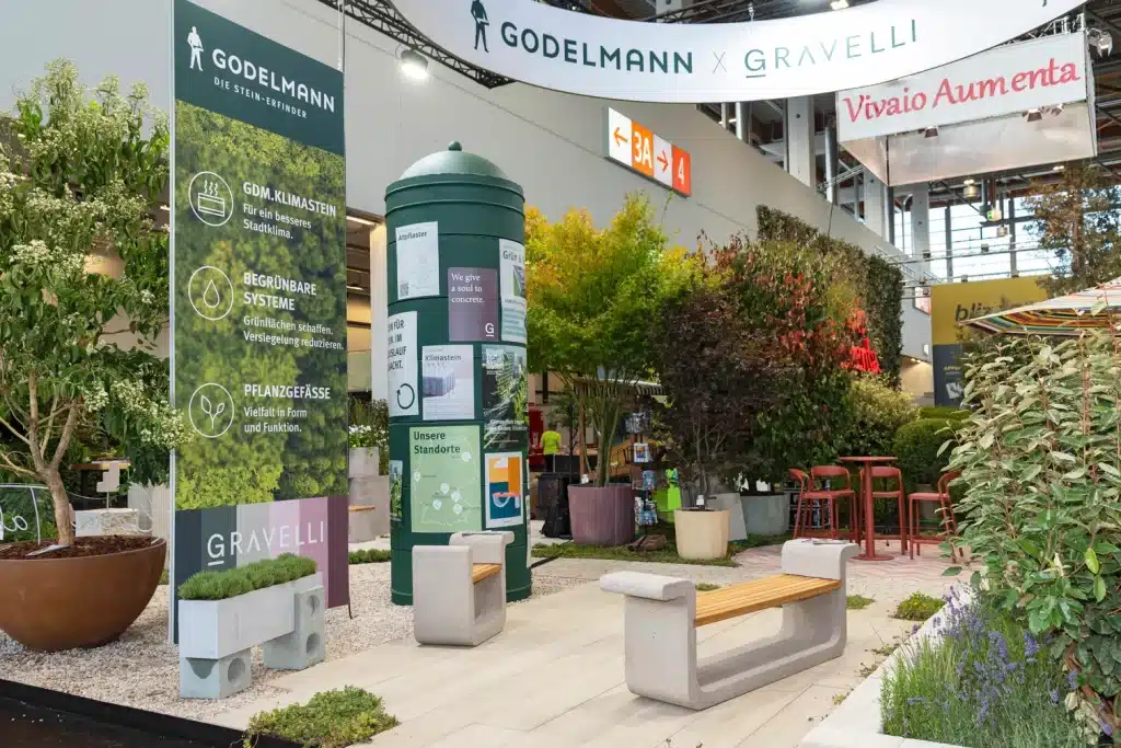 Godelmann-Stand auf der GaLaBau Nürnberg mit Gravelli-Banner, grüner Infosäule, Betonbänken und bepflanzten Gefäßen.