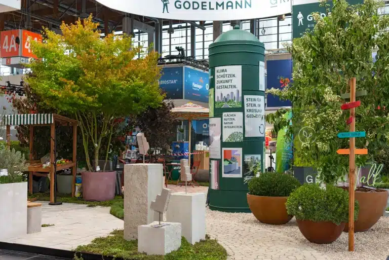 Detail des Godelmann-Messestands auf der GaLaBau Nürnberg mit grüner Infosäule, hellen Steinquadern und Wegweiser zwischen Pflanzinseln.