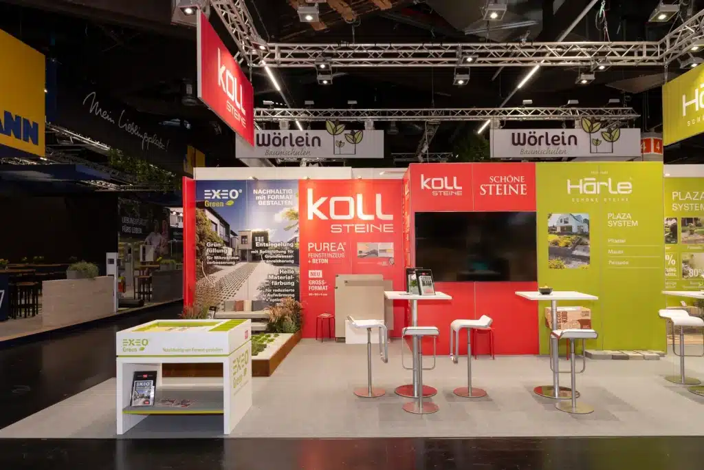 Frontansicht des KOLL-Messestands auf der GaLaBau Nürnberg mit roter Markenwand, Stehtischen, Barhockern und Infotheke EXEO Green.