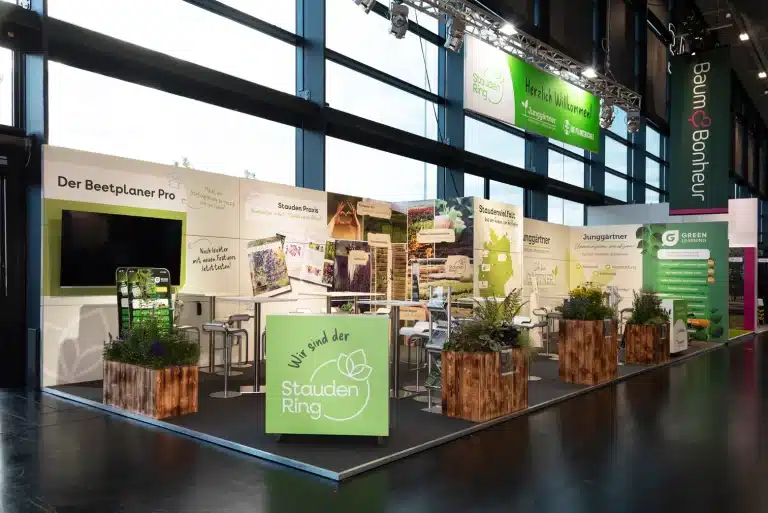 Messestand der Stauden Ring GmbH auf der GaLaBau in Nürnberg mit grüner Logowand, Holzpflanzkübeln und Würfel mit Schriftzug Wir sind der StaudenRing.