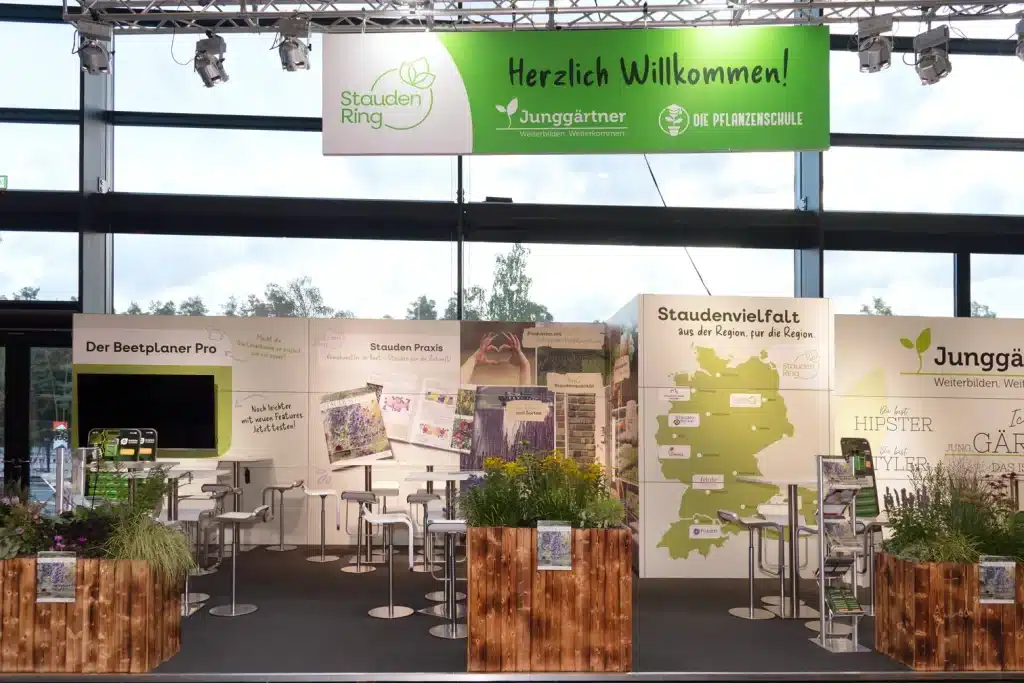 Frontansicht des StaudenRing-Messestands auf der GaLaBau Nürnberg mit Banner Herzlich Willkommen, Stehtischen und Deutschlandkarte Staudenvielfalt.