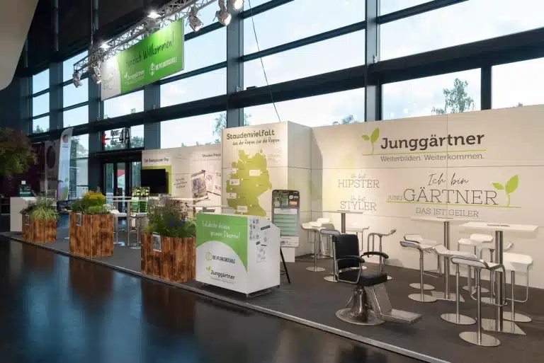 Messebau für den StaudenRing auf der GaLaBau in Nürnberg mit Junggärtner-Wand, Barhockern und bepflanzten Holzkübeln entlang der Standkante.