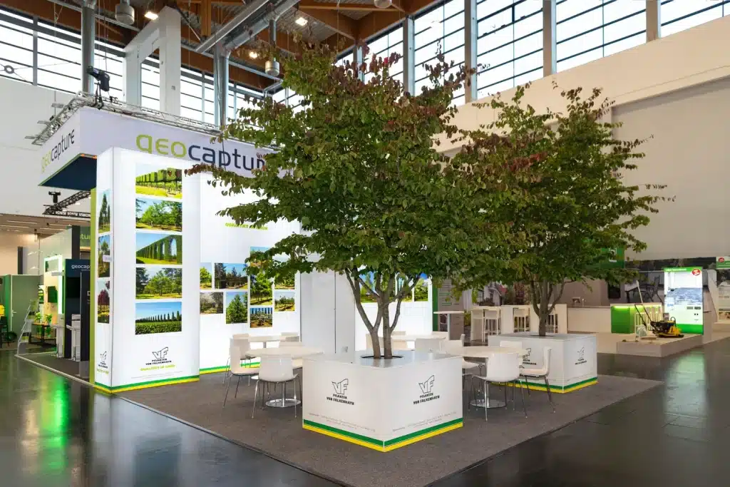 Messestand von VF-Pflanzen von Falkenhayn auf der GaLaBau in Nürnberg mit Bildwand, weißen Sitzgruppen und zwei großen Bäumen im Stand.
